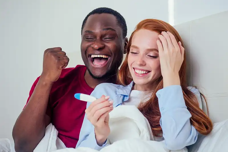 Feliz pareja de raza mixta sonriendo ante una prueba de embarazo positiva después de una fertilización in vitro.