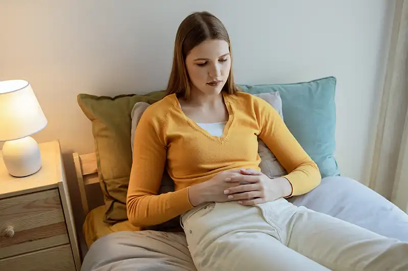 Mujer joven, sentada en una cama, luciendo pensativa mientras sostiene su estómago debido a endometriosis o dolor de síndrome de ovario poliquístico